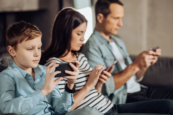 Kind, Frau und Mann sitzen nebeneinander auf einem Sofa und bedienen Smartphones