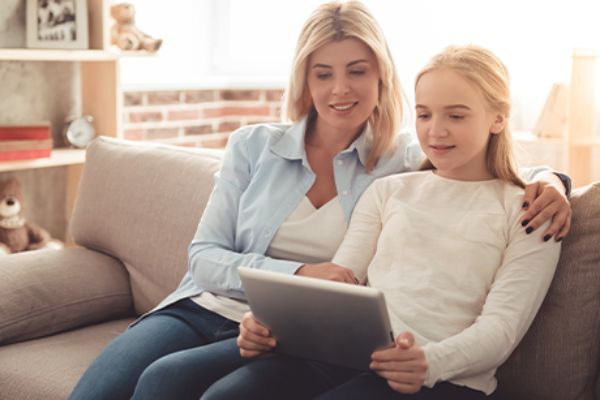 Mutter sitzt neben Mädchen mit Tablet auf einem Sofa.