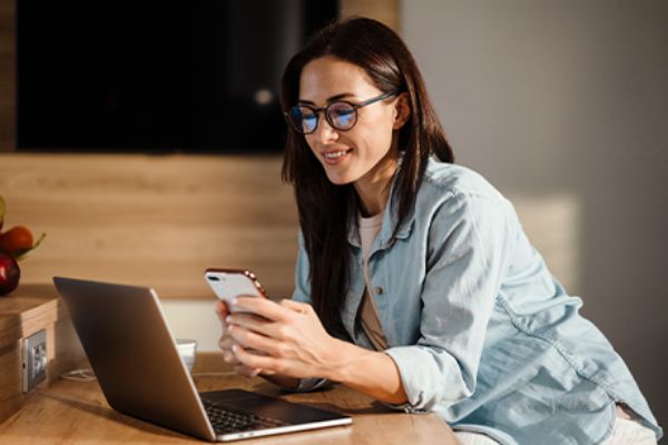 Frau sitzt lächelnd vor Laptop mit Smartphone in der Hand.