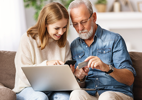 eine junge Frau und ein älterer Herr sitzen gemeinsam auf der Couch und schauen auf den Laptop und die Kreditkarte