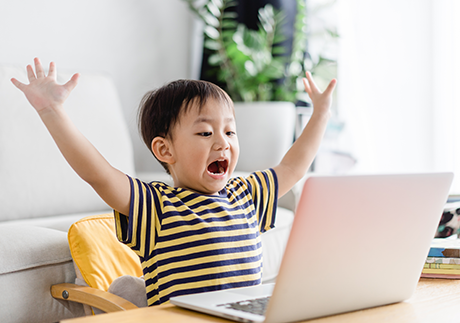 Kind sitzt vor dem Laptop und hebt die Arme in die Höhe.