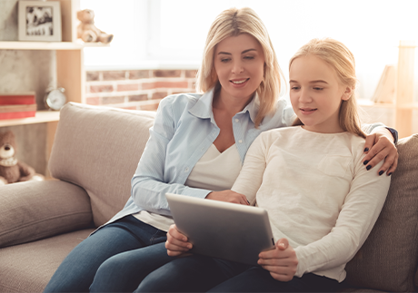 Mutter sitzt neben Mädchen mit Tablet auf einem Sofa.