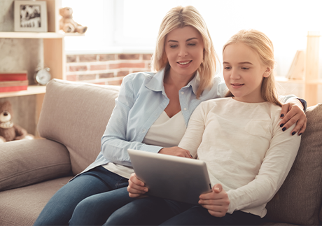 Mutter sitzt neben Mädchen mit Tablet auf einem Sofa. 